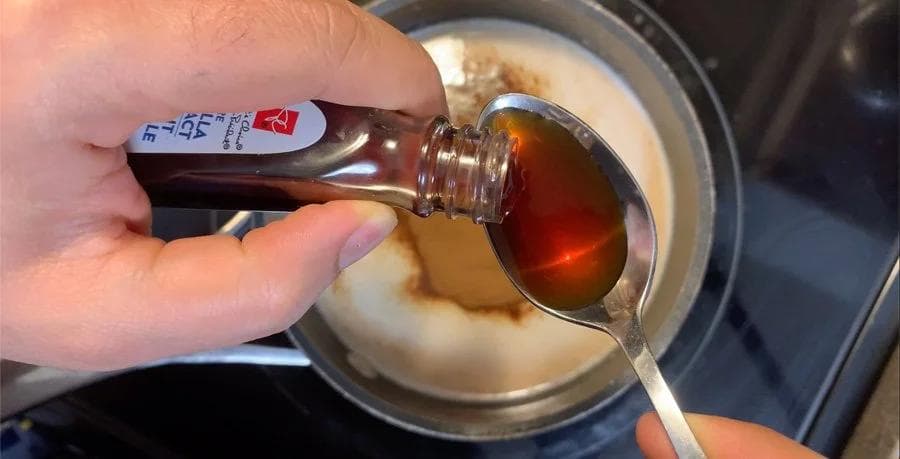 Pouring dark liquid coffee syrup from a bottle into a pot of cream-colored mocha mixture with a spoon.