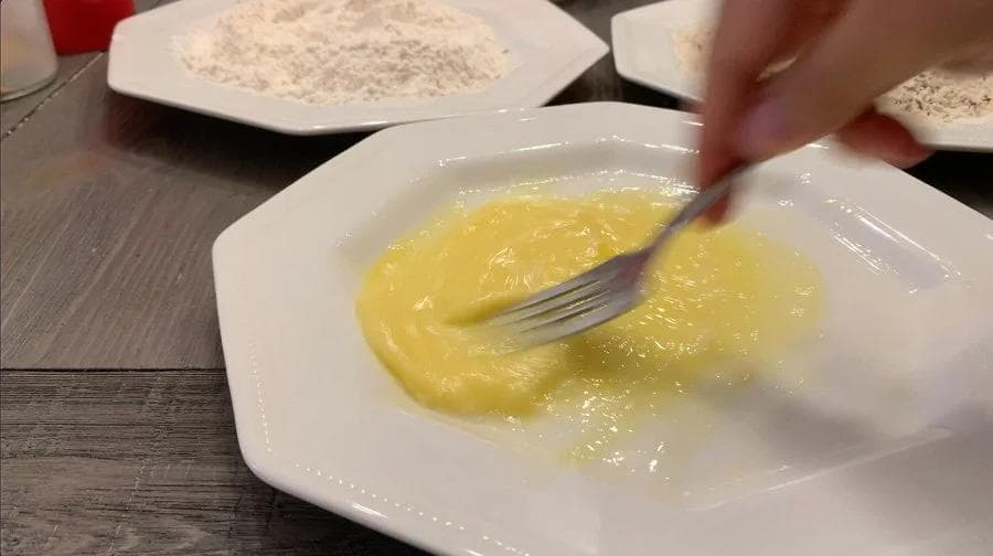 Hand whisking egg mixture in a white bowl during preparation of fried mozzarella balls breading station.