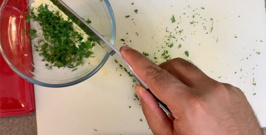 Hand spooning chopped fresh parsley into a bowl of creamy garlic butter mixture.