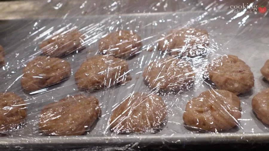 Shaped chicken nugget patties wrapped in plastic wrap on a baking tray, ready for freezing.