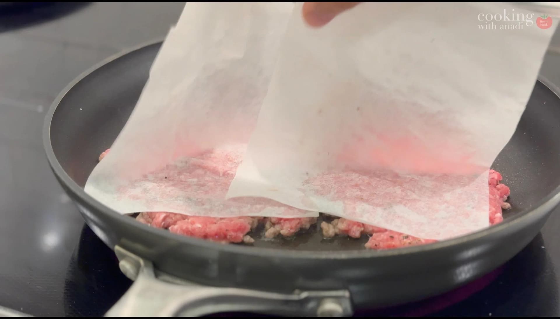 Parchment paper being peeled away from flattened burger patties with sliced onions in a skillet.