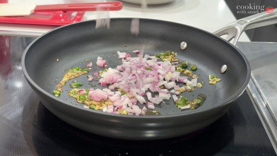 Sautéing diced pink onions with cumin seeds, green chiles, and ginger in a black wok on stovetop.