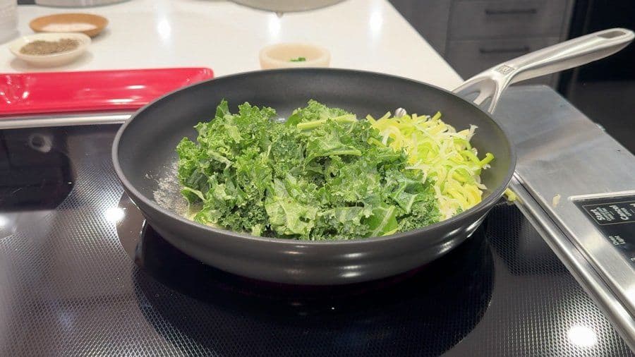 Sautéed leeks and kale wilting in a black pan with garlic-chive butter on a stovetop.