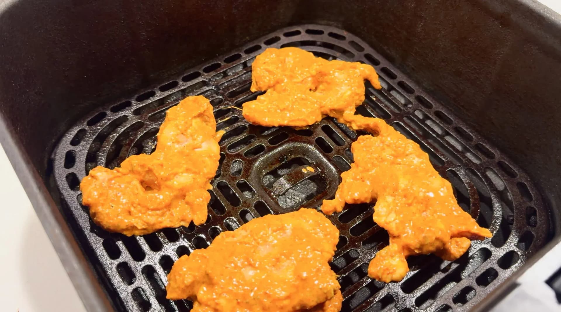 Marinated chicken tikka pieces coated in orange spiced yogurt arranged in a single layer on an air fryer rack.