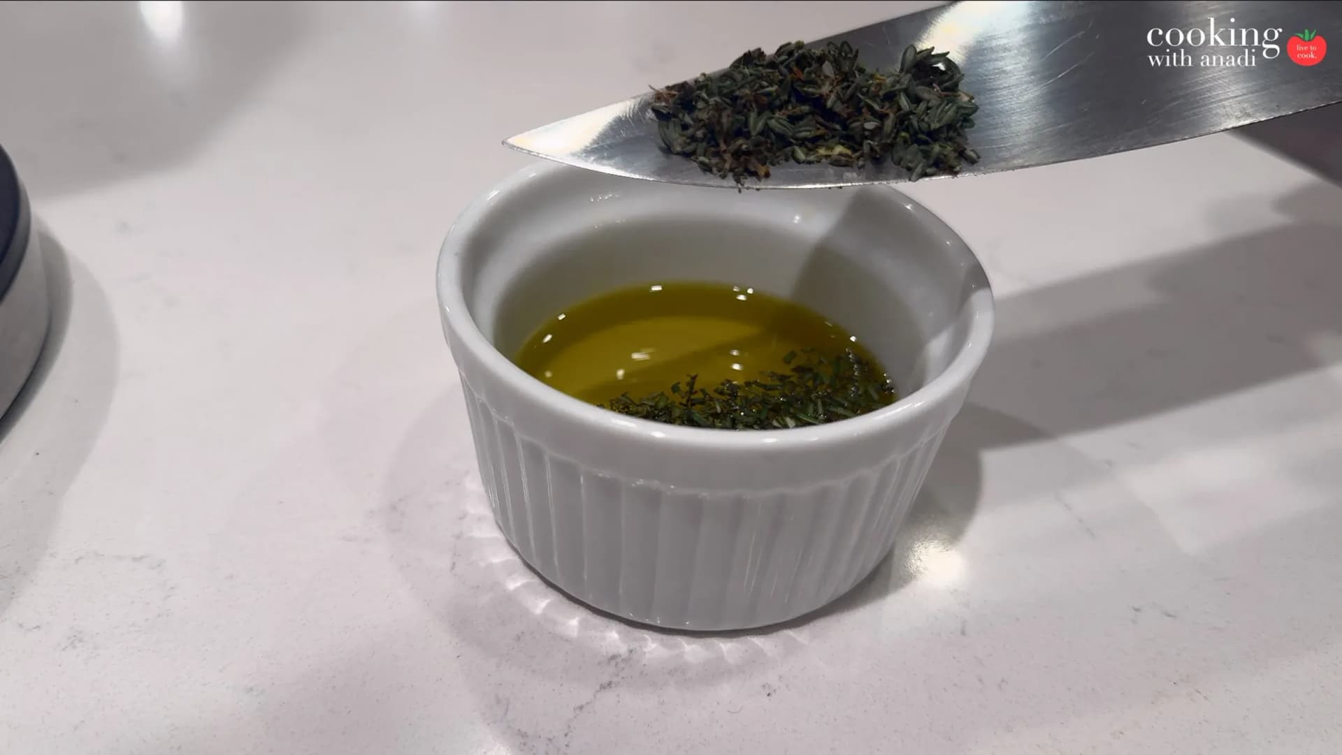Dried herbs being scraped from a knife blade into a small white ramekin filled with olive oil.