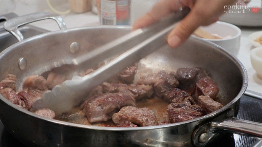 Searing beef cubes in a pan with tongs until lightly browned for Balinese beef rendang