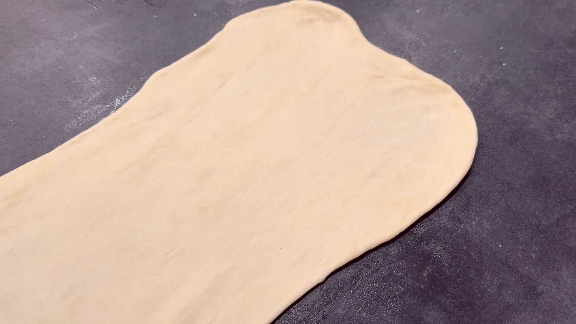 Thin, rolled-out Flammkuchen dough on a dark work surface, ready to be topped.