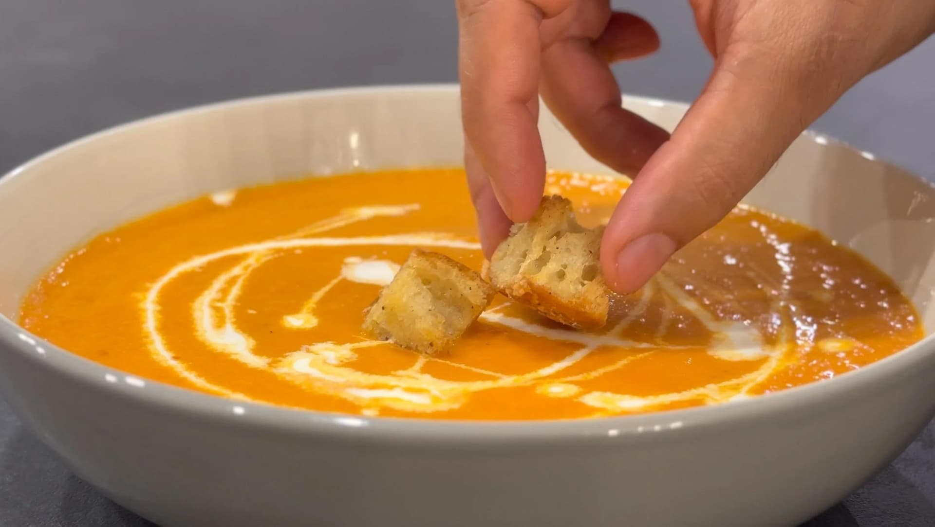 Creamy roasted tomato soup garnished with a swirl of cream and croutons being dipped into the bowl.
