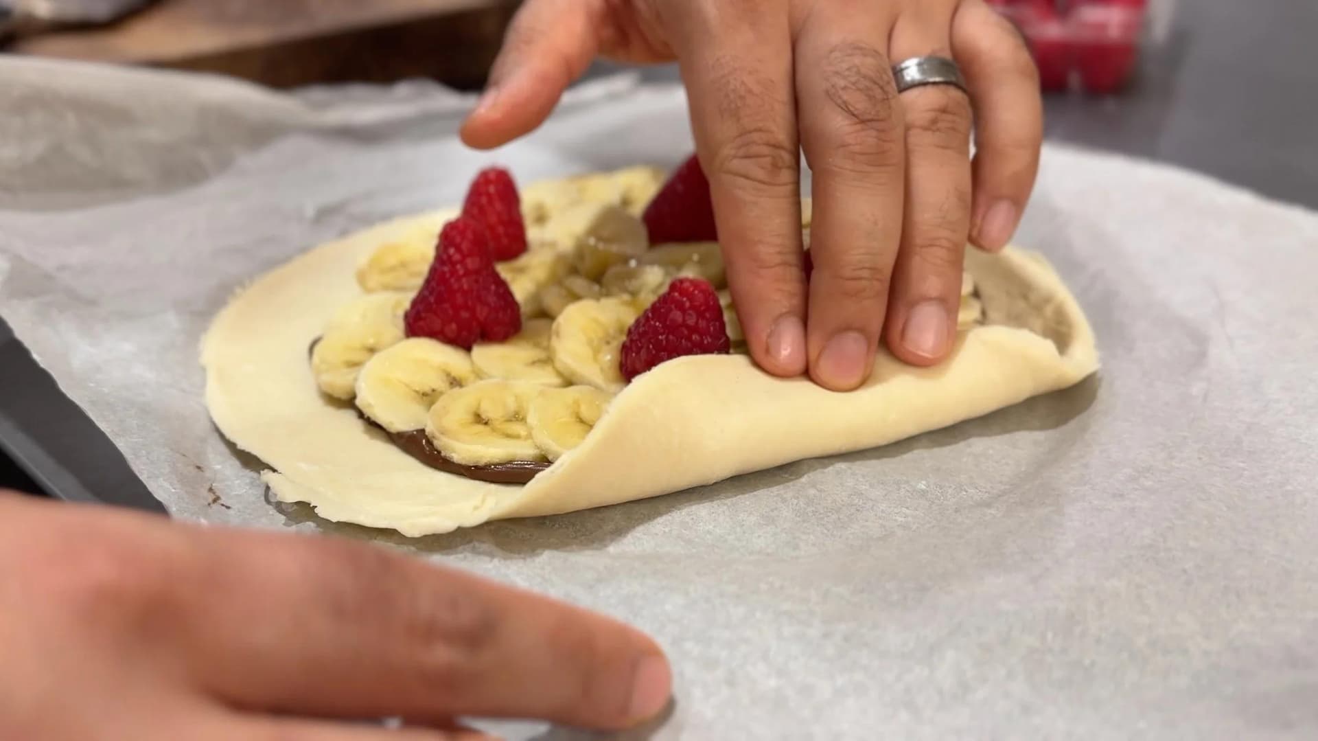 Hands folding the crimped edge of a banana Nutella galette with raspberries on top.
