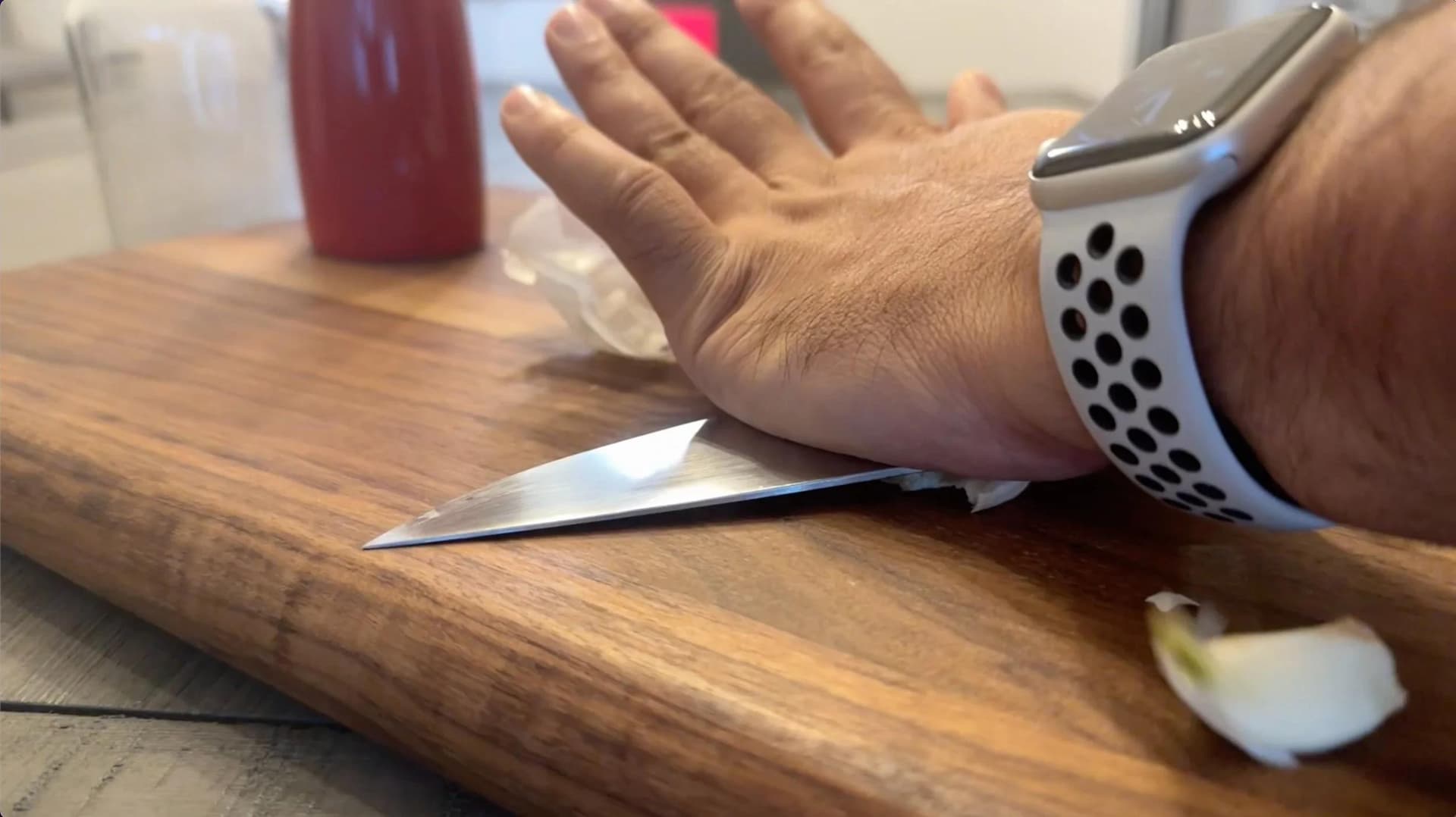 Hand using chef's knife to smash garlic clove on wooden cutting board.