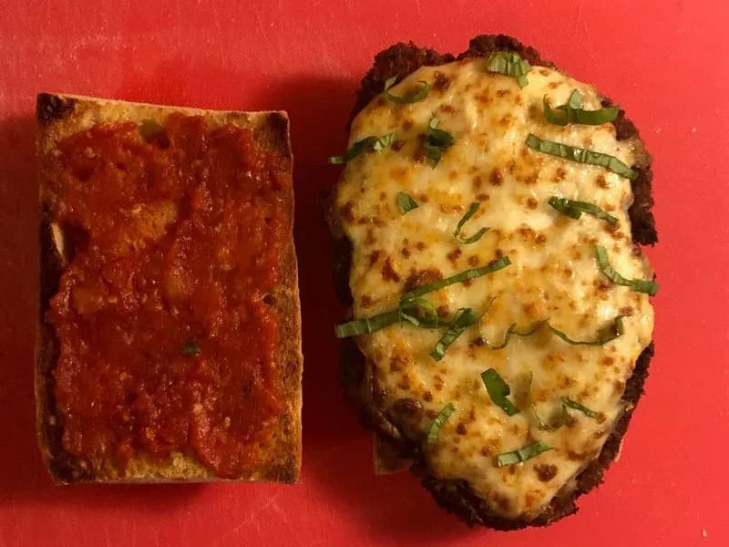 Two halves of a crispy chicken parmesan sandwich on ciabatta bread with melted cheese and fresh chives, alongside marinara sauce.