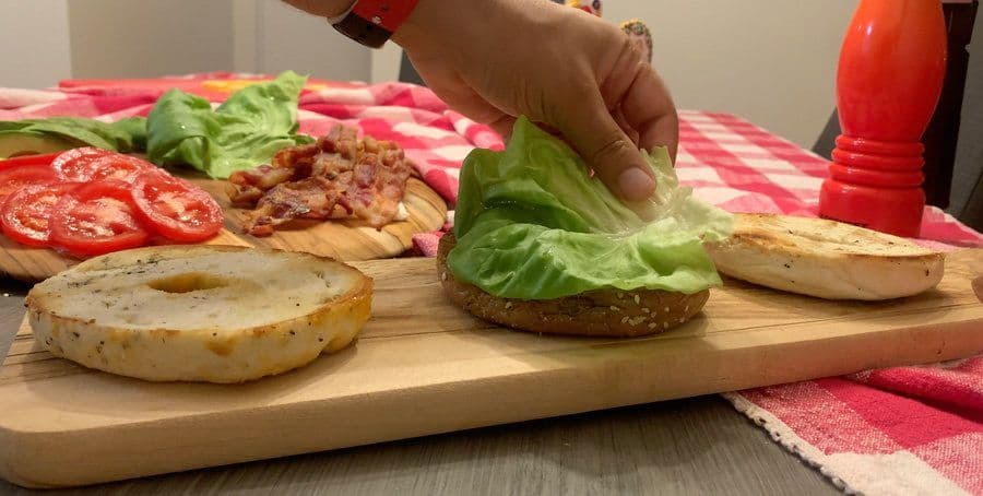 Hand assembling bagel sandwich with lettuce, tomato slices, bacon, and egg on wooden board.
