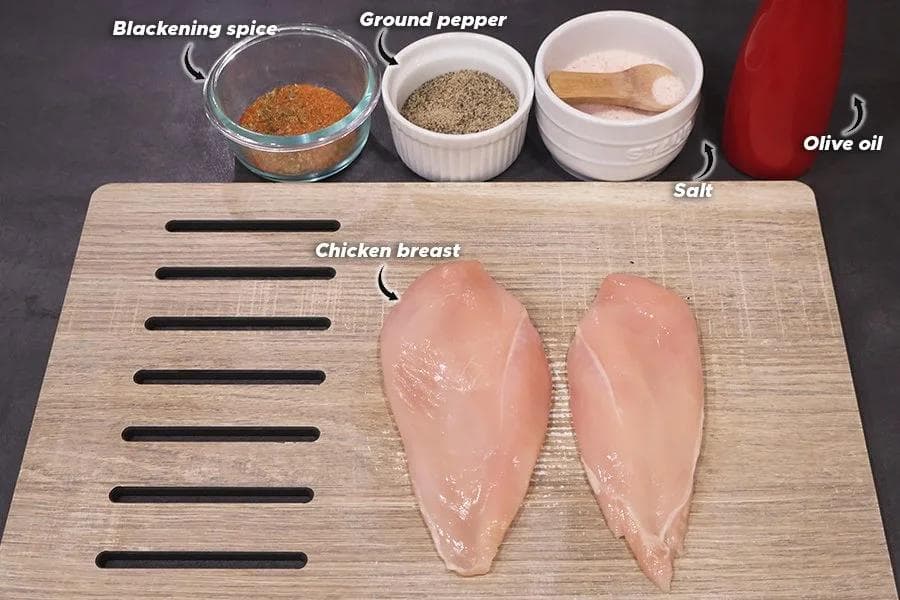 Ingredients for air fryer chicken breast laid out on a cutting board with spices, salt, olive oil, and two raw chicken breasts.