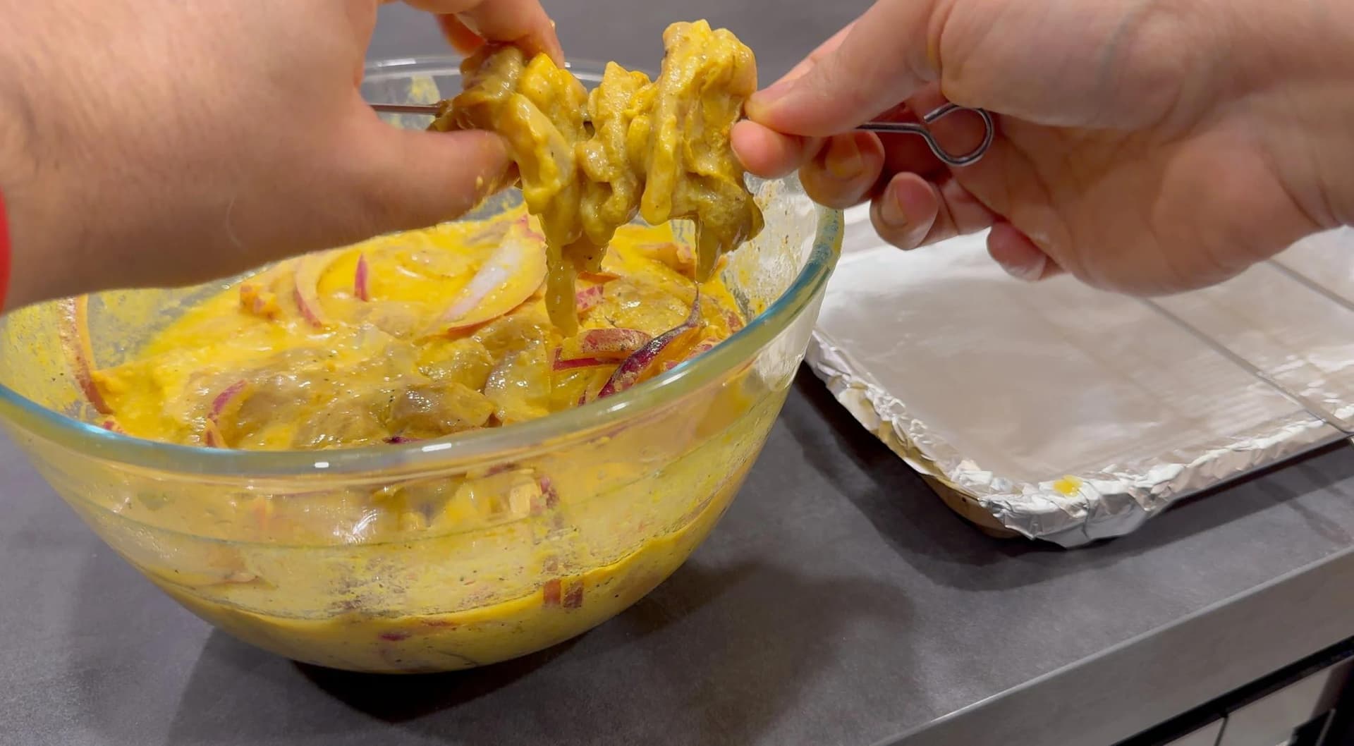 Hands lifting marinated chicken skewers from a glass bowl with yellow turmeric marinade.