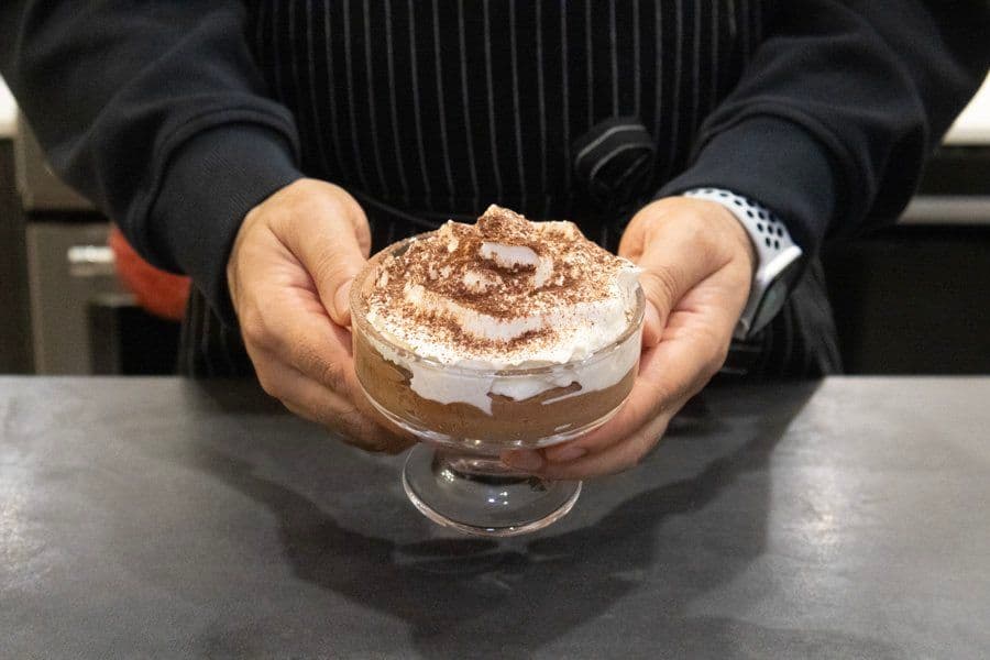 Chocolate mousse dessert in a glass cup topped with whipped cream and cocoa powder, held in hands.
