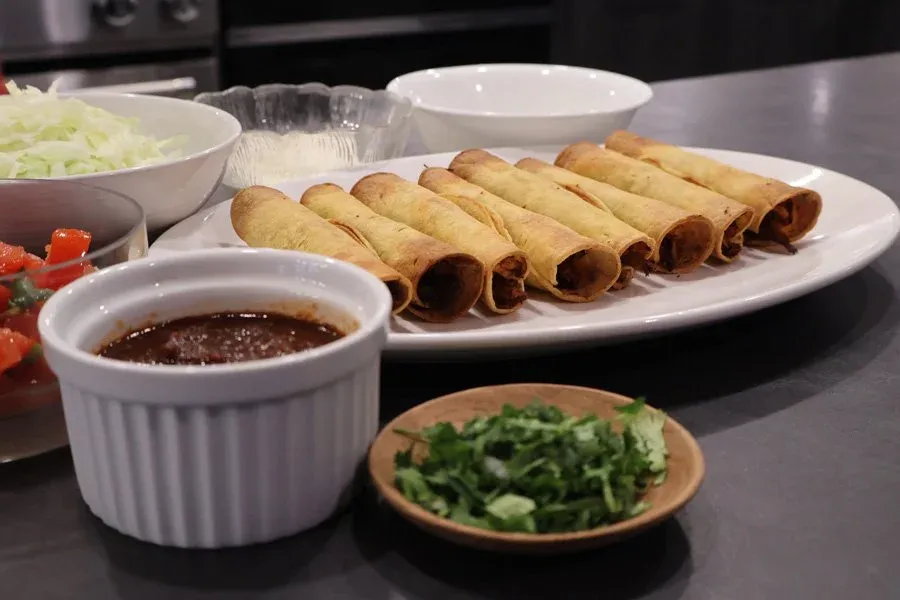 Crispy baked taquitos on a white platter with salsa roja dip, fresh cilantro, shredded lettuce, and diced tomatoes.