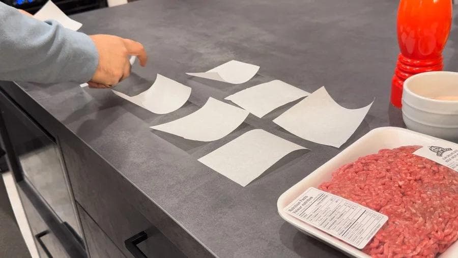 Twelve portioned ground beef balls on parchment paper squares with raw ground beef on a plate, prepared for smash burger sliders.