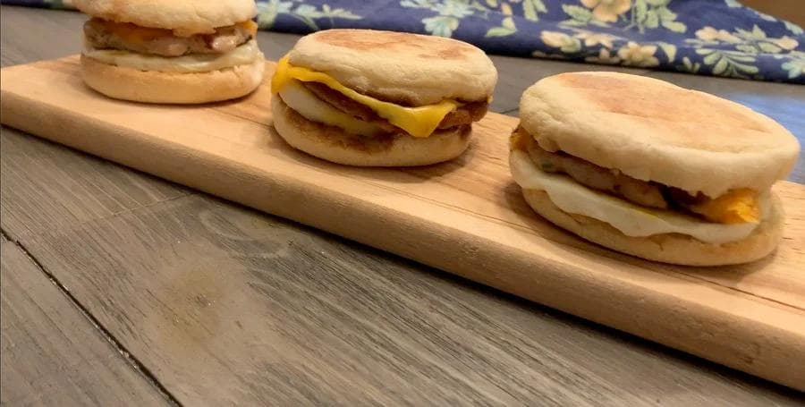 Three homemade breakfast sandwiches on a wooden board with English muffins, sausage patties, cheese, and egg.