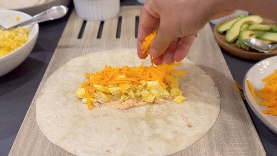 Hand adding shredded cheddar cheese to a breakfast burrito with eggs and ham on a flour tortilla.