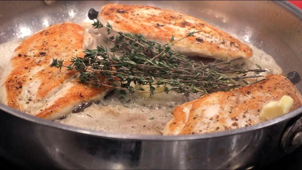 Pan-fried chicken breasts in butter with fresh thyme sprigs and garlic cloves in a stainless steel skillet.
