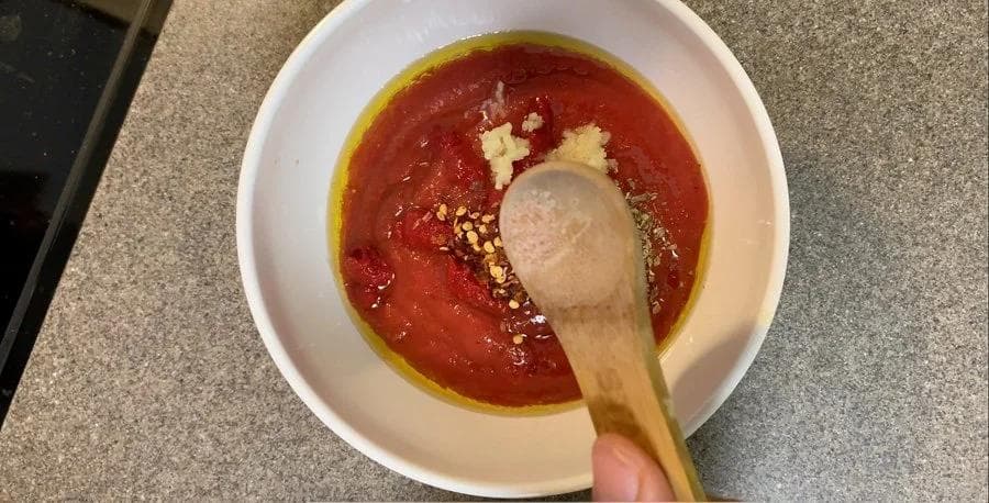 Pizza sauce ingredients combined in a white bowl with a wooden spoon, showing red tomato base with minced garlic.