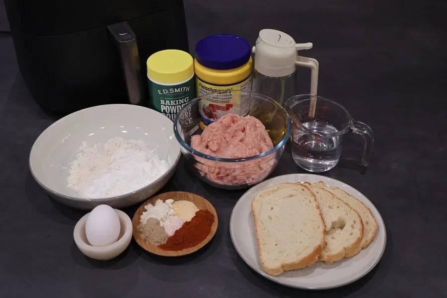 Ingredients for chicken nuggets laid out including ground chicken, bread slices, egg, flour, spices, and condiment bottles.