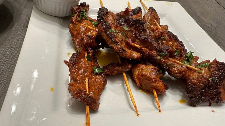 Grilled beef skewers on wooden sticks arranged on a white plate with a small bowl of sauce in the background.