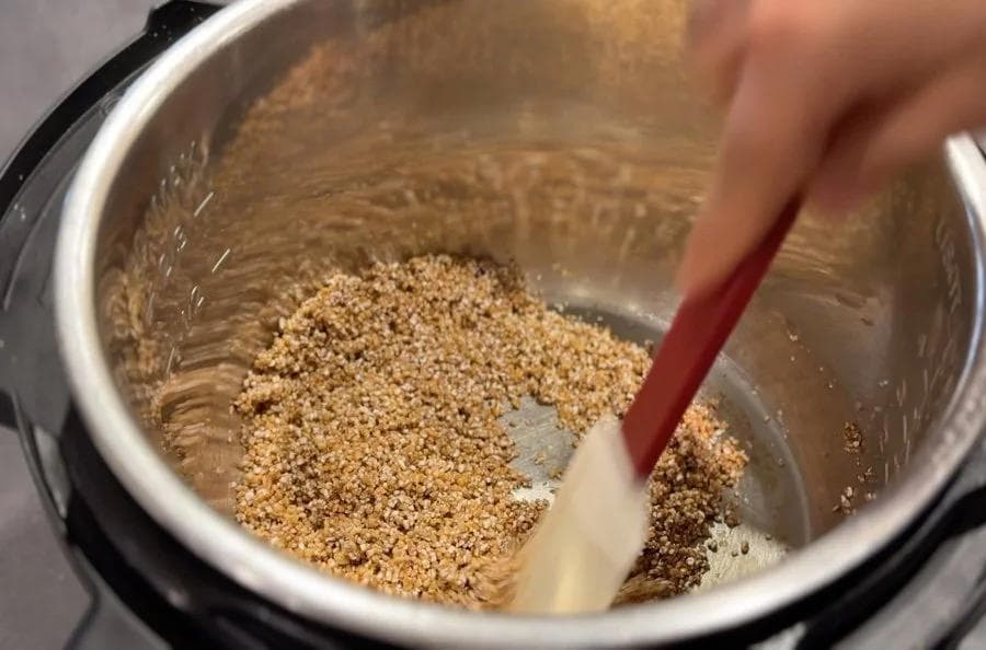 Roasting cracked wheat in ghee in an Instant Pot using a spatula, grains turning golden brown.