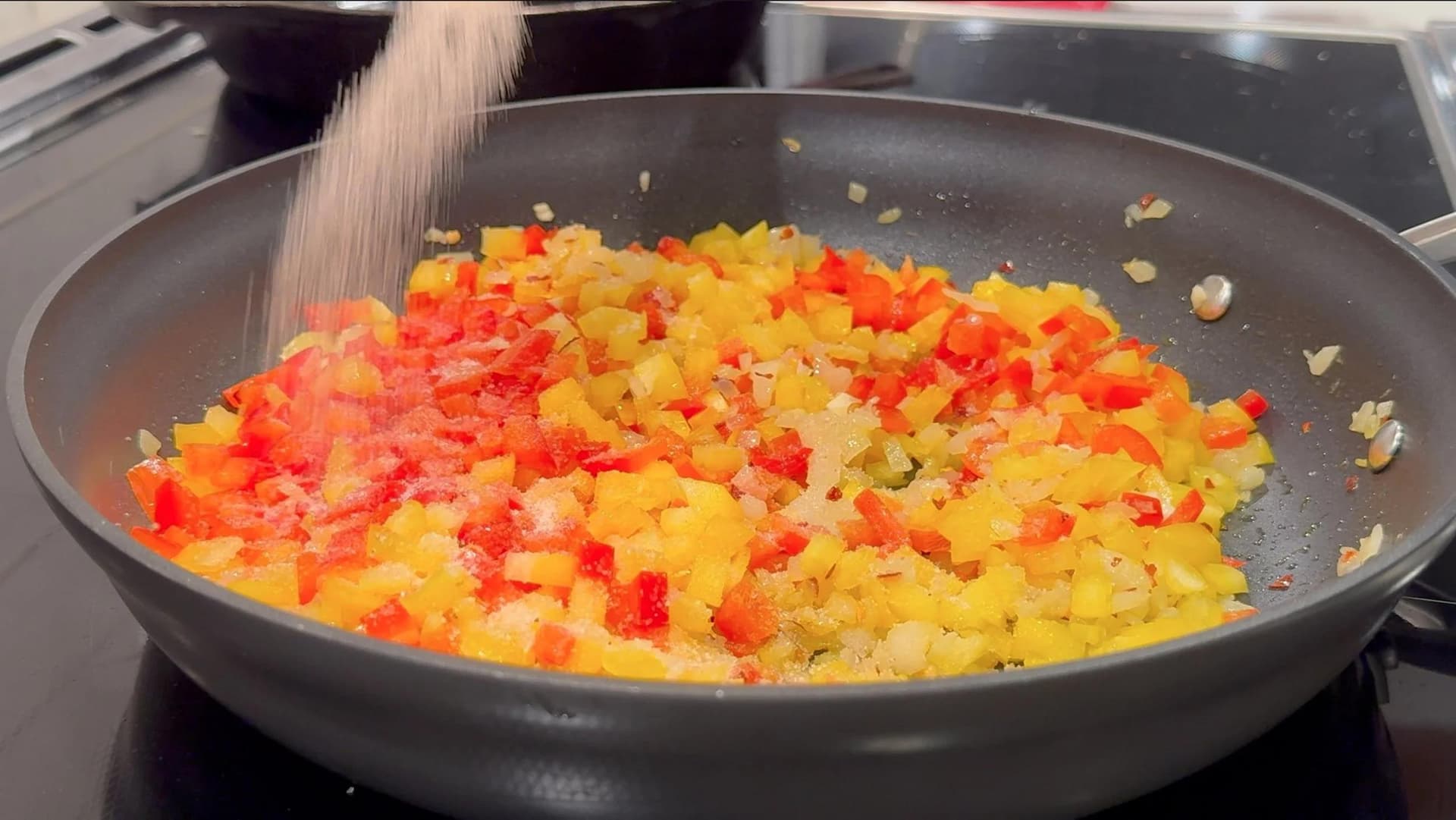 Salt being sprinkled over diced red and yellow bell peppers with onion sautéing in a large black skillet.