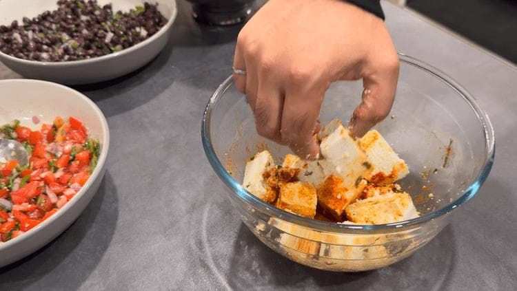Hand mixing marinated paneer cubes in a glass bowl during paneer pakora preparation.
