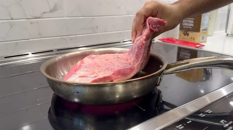 Hand lifting seared pink meat from a stainless steel pan on a stovetop during birria preparation.