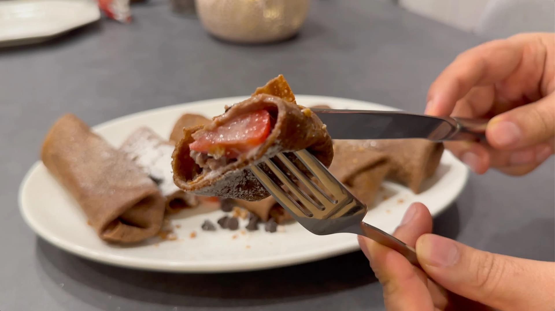 Chocolate crepe being cut with a fork, revealing red jam filling and melted chocolate interior on a white plate.