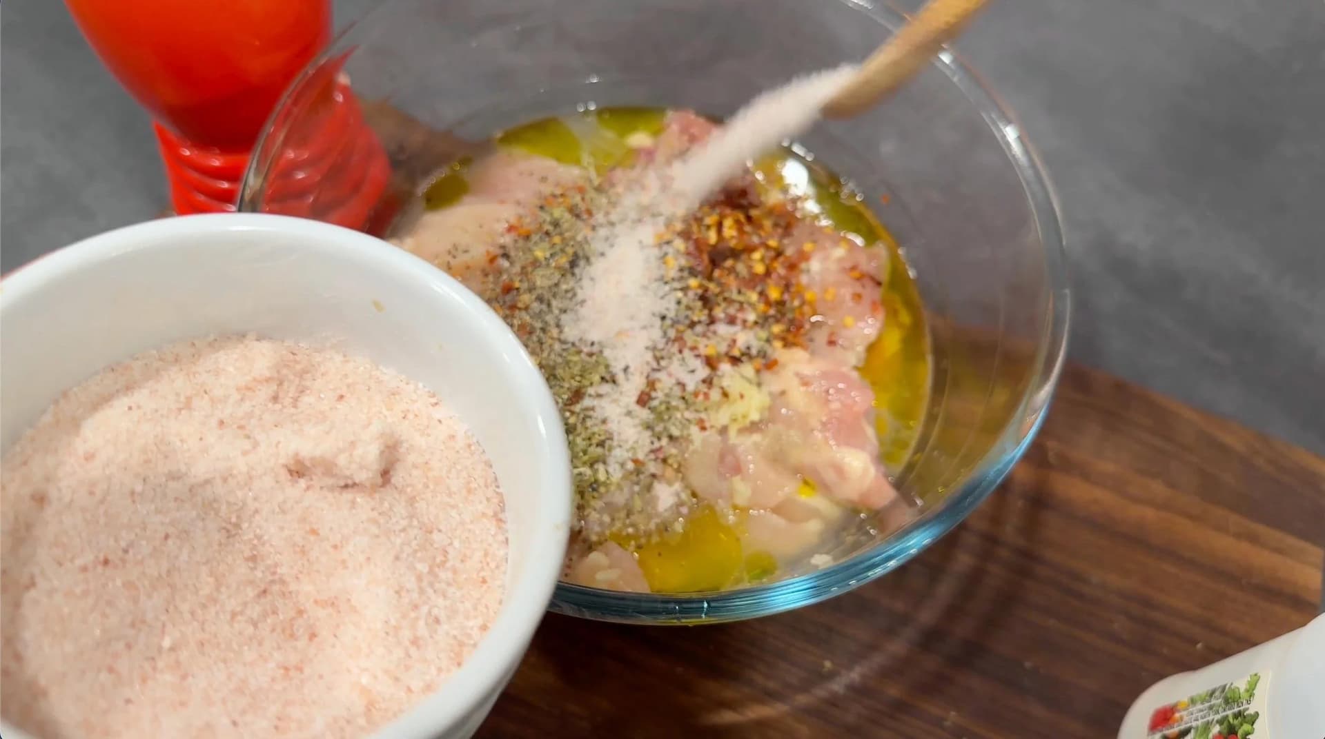 Seasoning being added to marinated chicken cubes in a glass bowl with olive oil and herbs for souvlaki marinade.