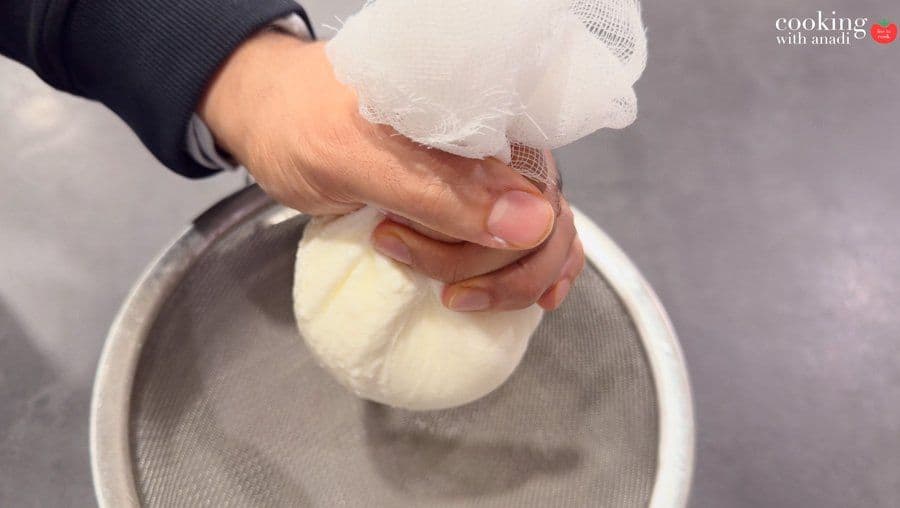 Hands squeezing a cheesecloth bundle of yogurt over a fine mesh sieve to drain excess moisture.
