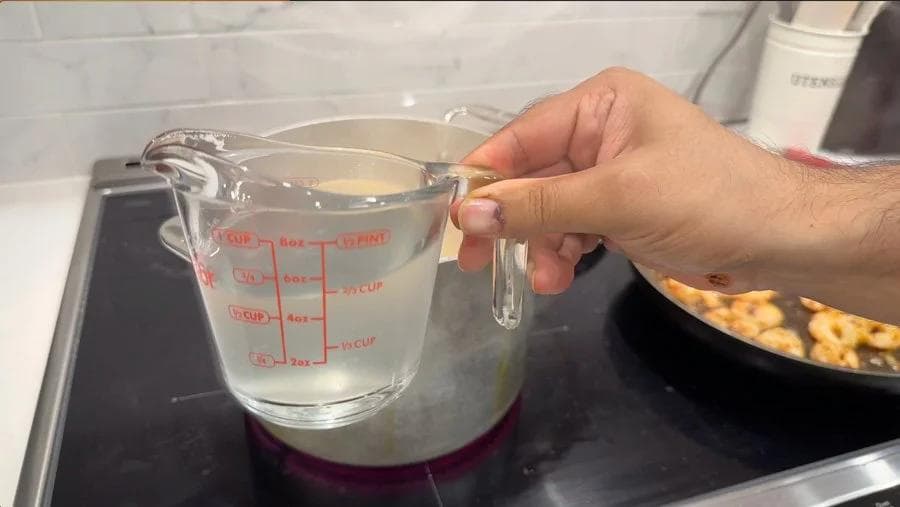 Hand holding a measuring cup of reserved pasta water on a stovetop during pasta preparation.
