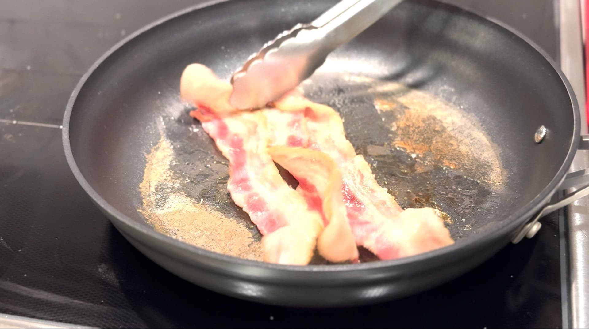Bacon strips cooking in a skillet with water, being turned with a spatula as fat renders.