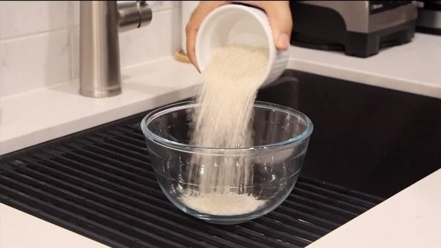 Sugar being poured into a glass bowl of cooked rice pudding mixture in a kitchen sink.