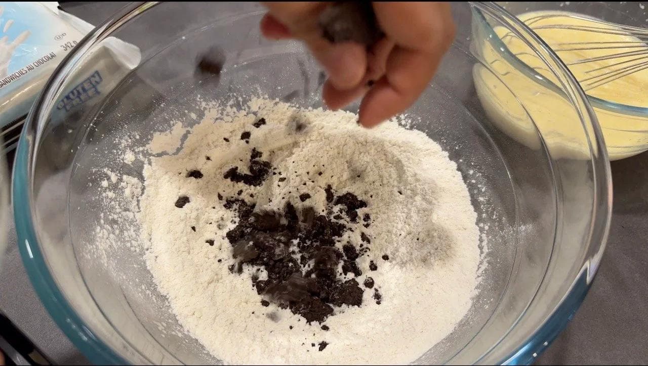 Crushed Oreo cookie pieces being added to white dry pancake batter in a glass bowl.