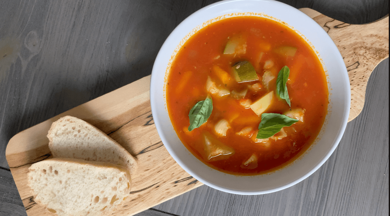 Bowl of vegan minestrone soup with vegetables and broth, garnished with basil, served with crusty bread.