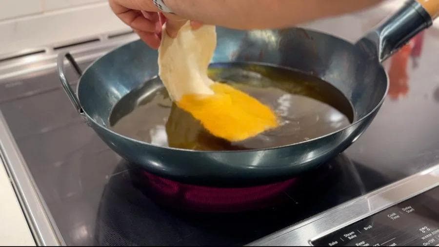 Hand placing a fried pastry dough into hot oil in a wok using a slotted spoon.