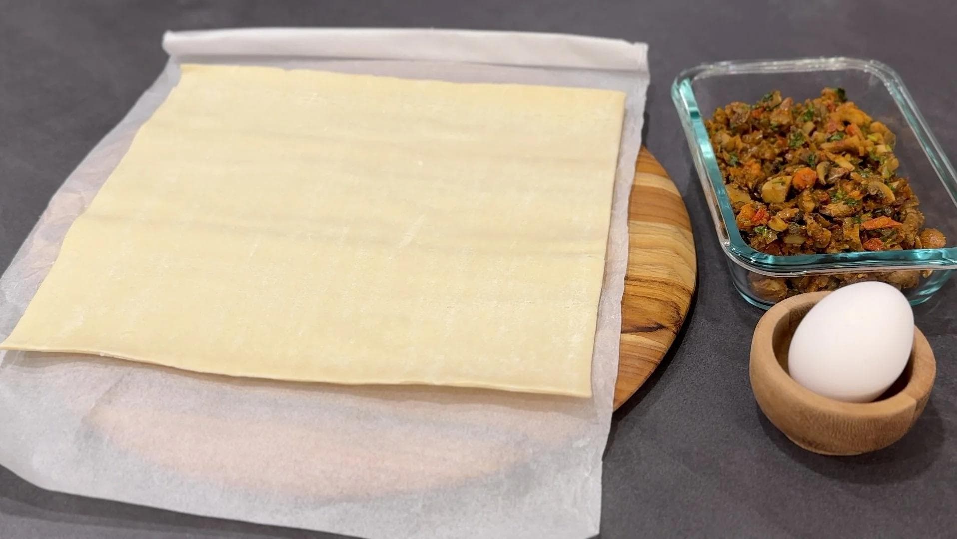 Puff pastry sheet on parchment paper with a bowl of mushroom filling and an egg beside it, ready for assembly.