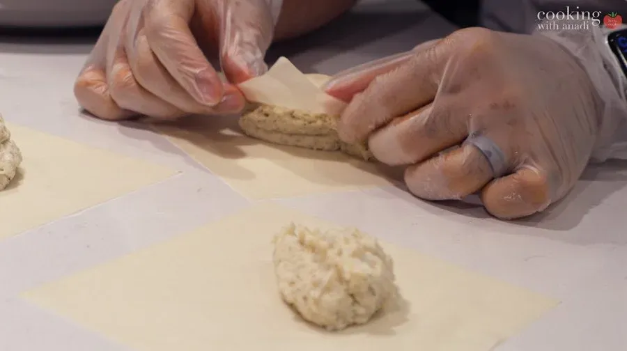 Hands folding a spring roll wrapper over mashed potato filling on a white surface.