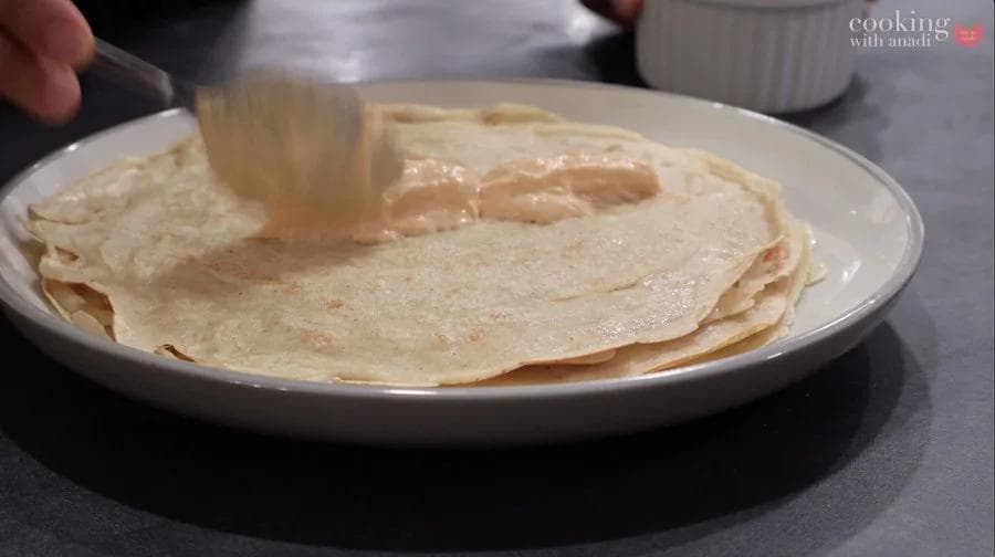 Cooked pumpkin crepes stacked on a white plate with butter being spread across the top one.