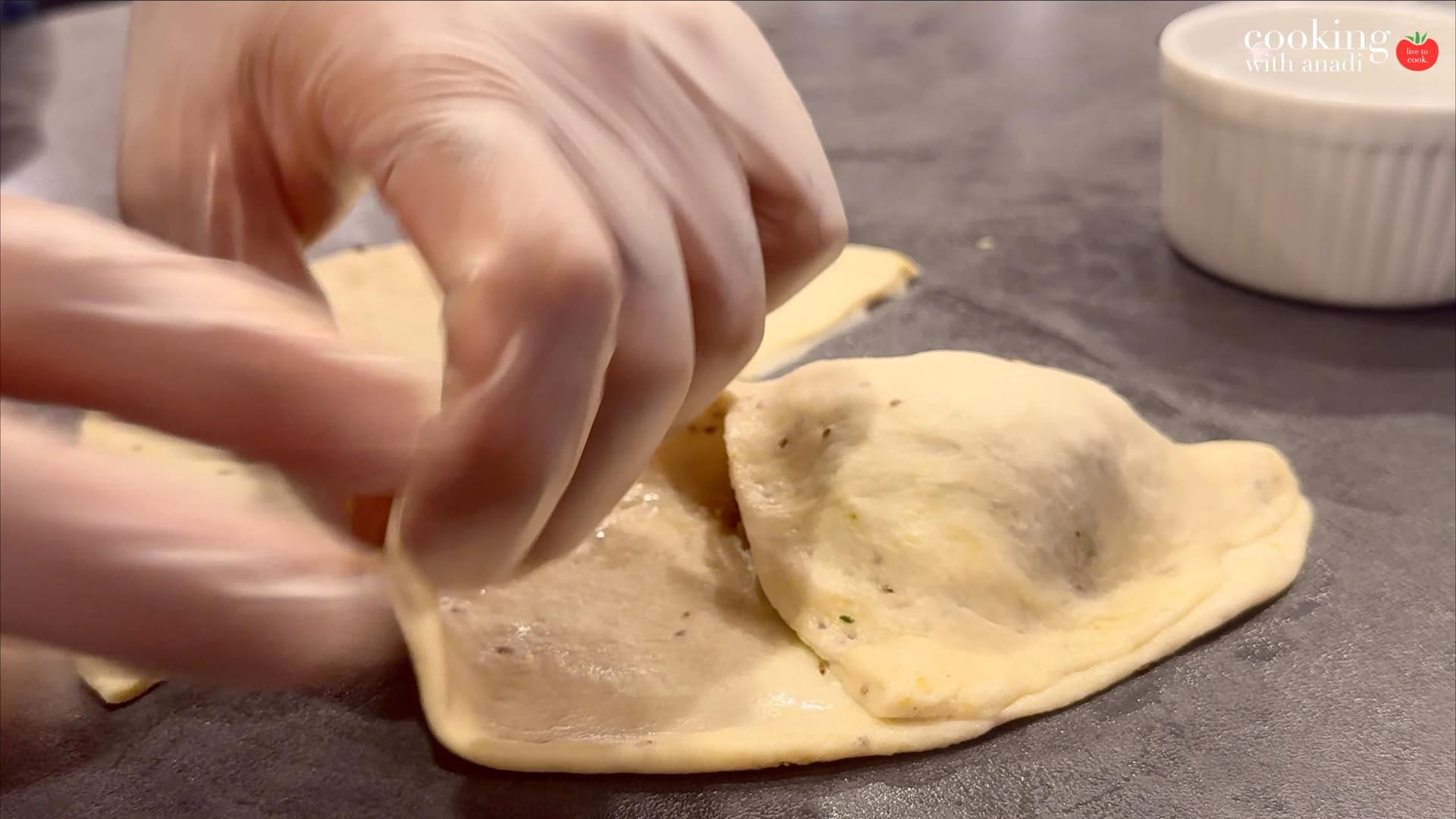 Hands folding and sealing a samosa by pinching the edges together on a dark work surface.