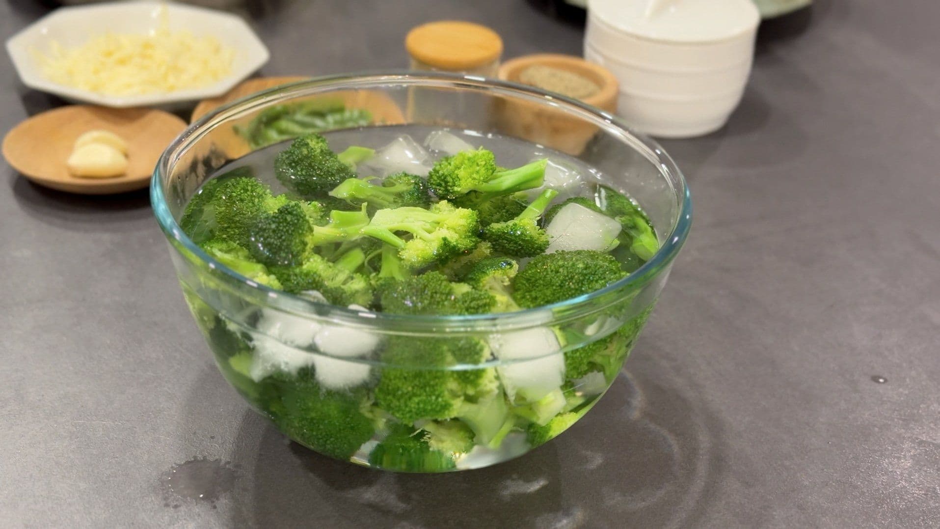 Blanched broccoli florets in ice water bath with ingredients arranged in bowls on a gray countertop.