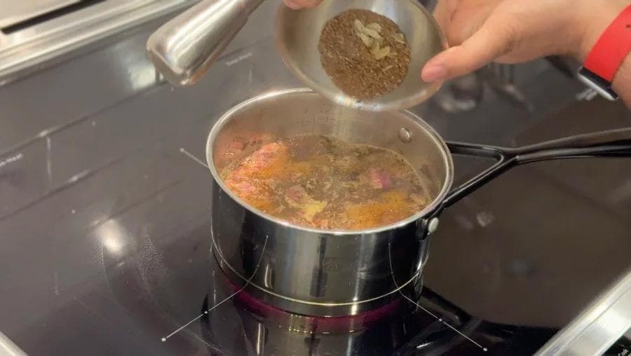 Pouring ground chai masala spices into a saucepan of simmering tea with rose petals and ginger.
