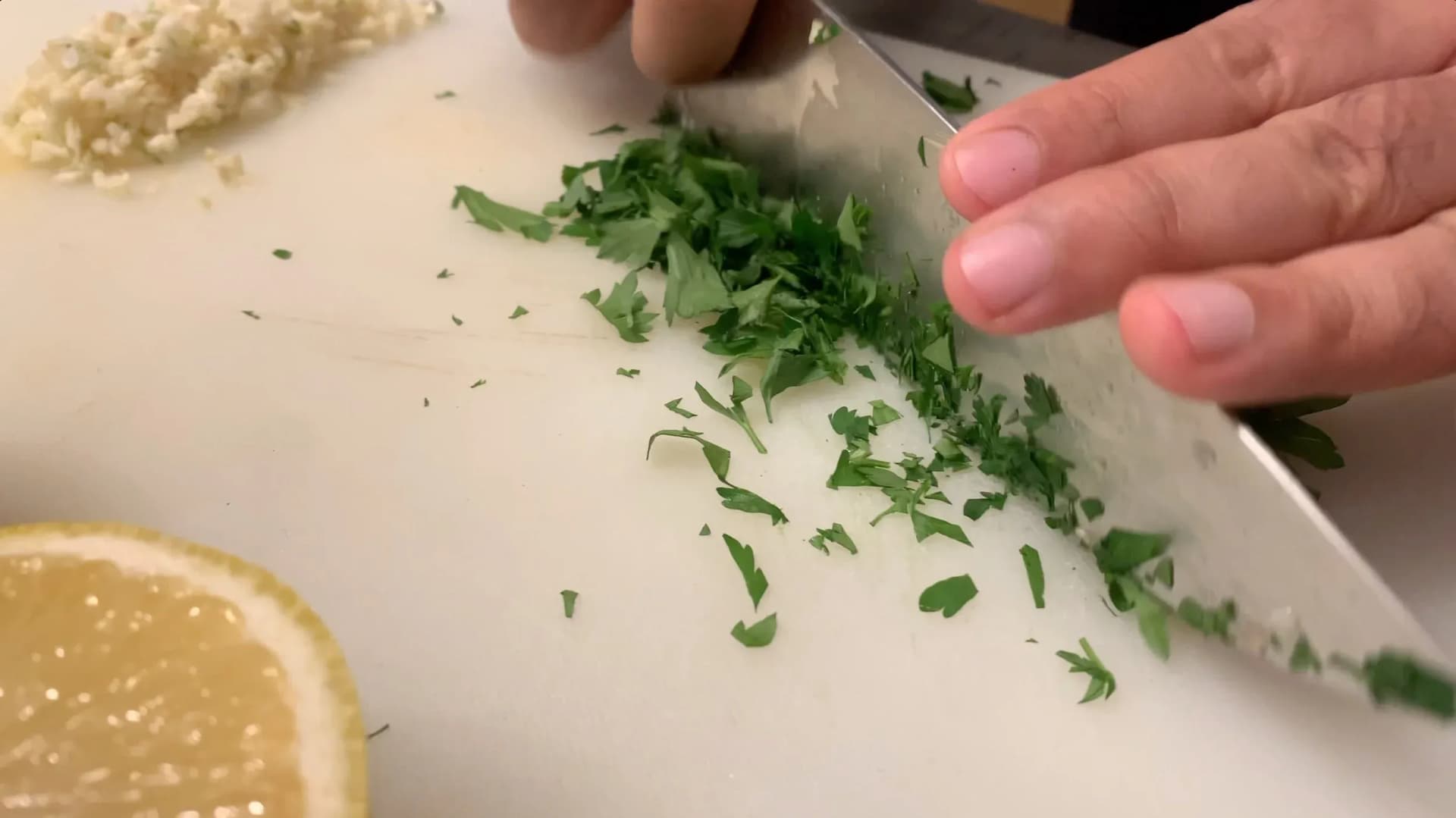 Chopped fresh parsley being added to shrimp scampi sauce on a cutting board with lemon and garlic nearby.