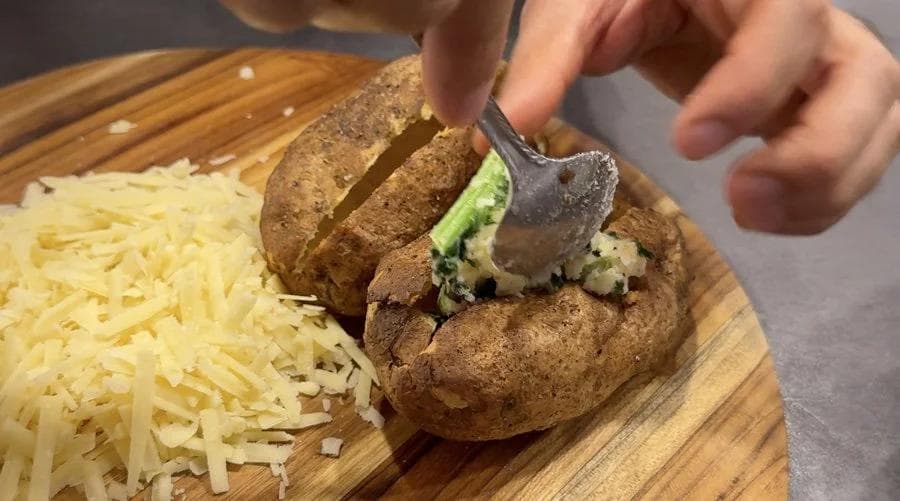 Spoon filling colcannon mixture being added back into halved potato shells on a wooden board.