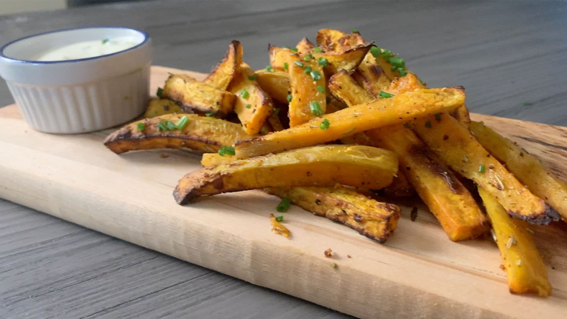 Crispy air fryer sweet potato fries on a wooden board with fresh herbs and a white dipping sauce.