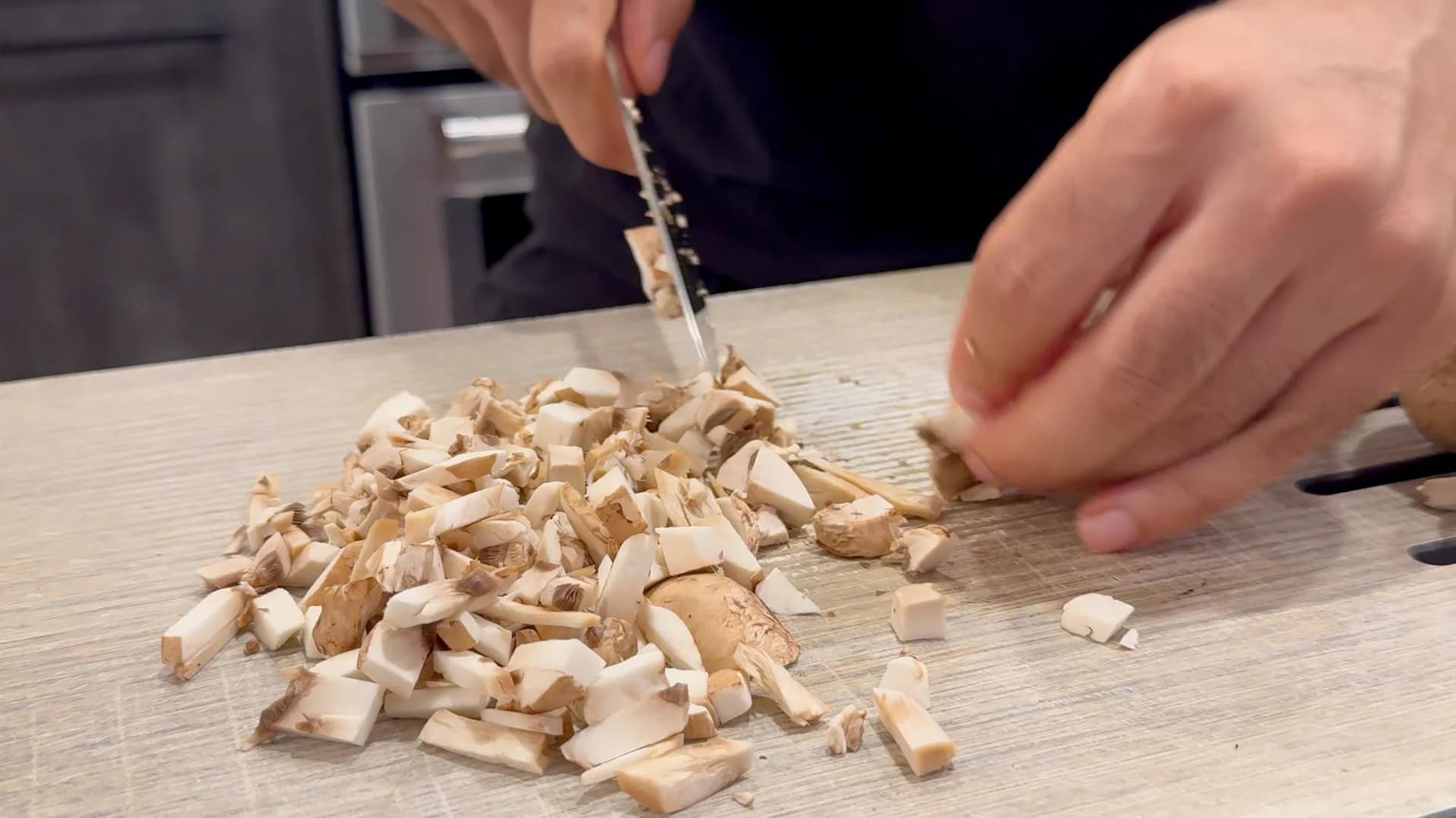 Chopped mushrooms being added to a wooden cutting board during preparation of Polish croquette filling.